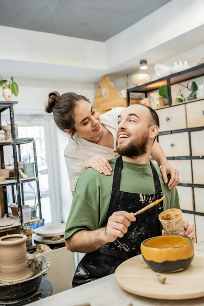 Craftswoman positivo no abraço avental e olhando para namorado colorir tigela de argila no estúdio de cerâmica — Fotografia de Stock