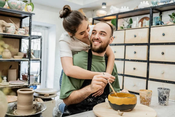 Joyful potter embracing boyfriend coloring clay bowl and working in ceramic studio at background — Stock Photo