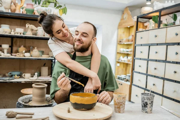 Smiling female potter hugging boyfriend coloring clay bowl while working in ceramic workshop — Stock Photo
