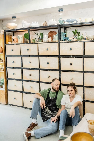 Craftsman in apron looking at smiling girlfriend sitting near cupboard in ceramic studio — Stock Photo