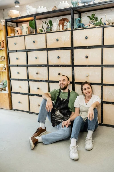 Couple agréable d'artisans dans des tabliers assis près de placard en atelier de céramique à l'arrière-plan — Photo de stock