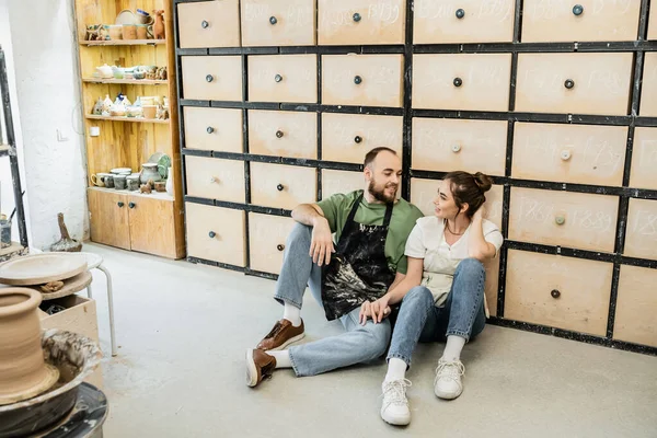Alegre pareja de alfareros en delantales tomados de la mano mientras están sentados cerca del armario en el estudio de cerámica - foto de stock