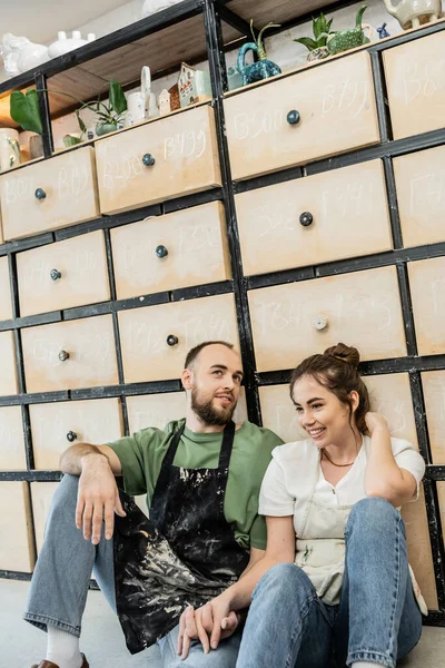 Happy couple of potters holding hands while sitting near cupboard in ceramic workshop — Stock Photo
