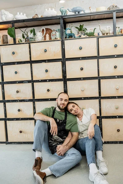Lächelndes Handwerkerpaar hält Händchen und blickt in die Kamera neben dem Schrank in der Keramikwerkstatt — Stockfoto