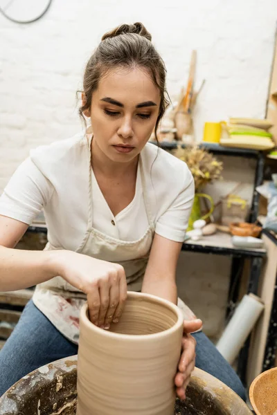 Brünette Handwerkerin in Schürze, die Ton auf Töpferscheibe im Keramik-Atelier im Hintergrund formt — Stockfoto