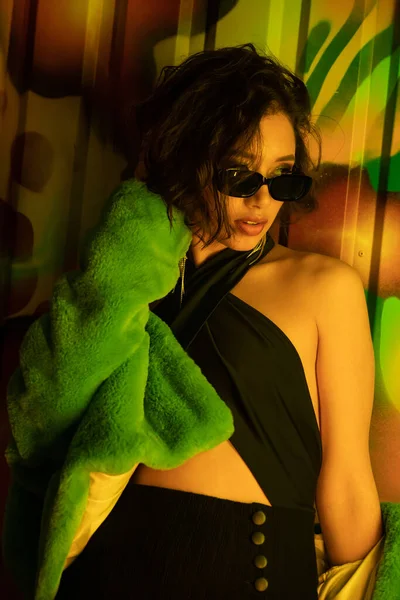Стильная молодая азиатка в солнцезащитных очках и искусственной меховой куртке, стоящая возле граффити в ночном клубе — стоковое фото