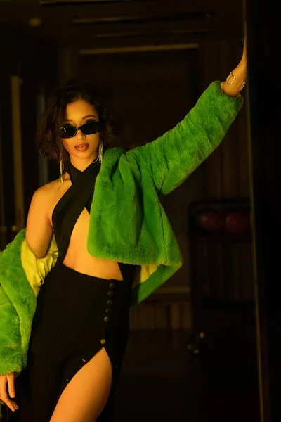 Модная молодая азиатская женщина в платье, солнцезащитных очках и искусственной меховой куртке позирует в темном ночном клубе — стоковое фото