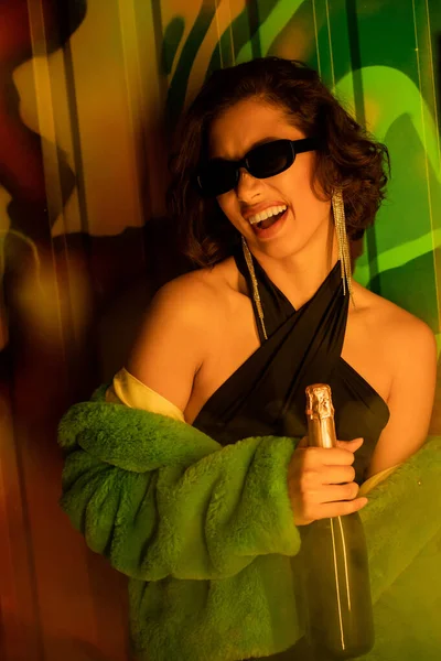 Emocionado mujer asiática en gafas de sol y chaqueta de piel falsa celebración de champán cerca de graffiti en el club nocturno - foto de stock