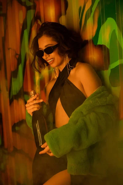Positive brunette woman in sunglasses and dress holding champagne near graffiti in night club — Stock Photo