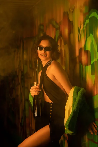 Fashionable young woman in sunglasses and dress holding champagne near graffiti in night club — Stock Photo
