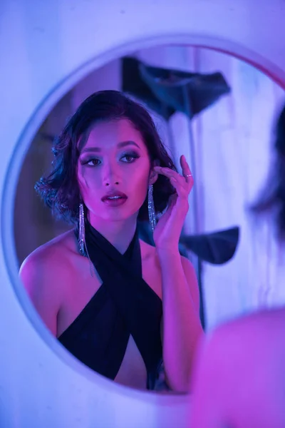 Elegante giovane bruna donna asiatica toccando orecchino vicino specchio e luce al neon nel night club — Foto stock