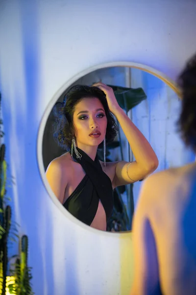 Jeune femme asiatique à la mode en robe touchant les cheveux près du miroir et de la lumière au néon dans une boîte de nuit — Photo de stock