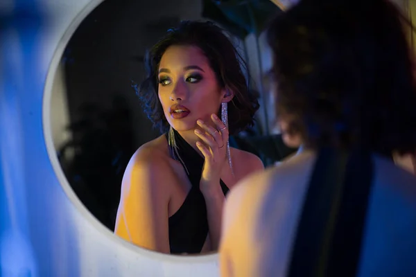 Модна молода азіатська жінка в одязі дивиться далеко від дзеркала в неоновому світлі в нічному клубі — стокове фото