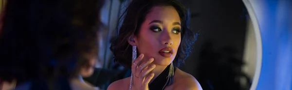 Elegante joven mujer asiática con maquillaje mirando lejos cerca del espejo y la luz de neón en el club nocturno, pancarta - foto de stock