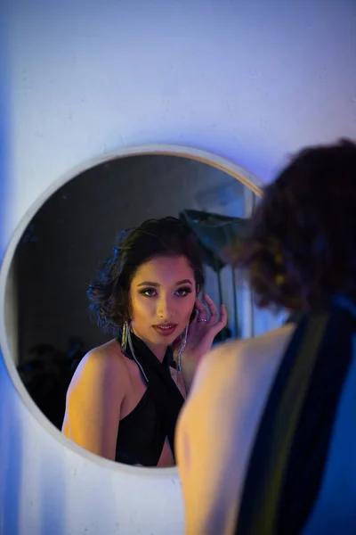 Femme asiatique confiante en tenue élégante et maquillage en regardant le miroir près de la lumière au néon dans la boîte de nuit — Photo de stock