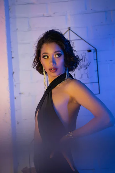 Femme asiatique à la mode avec maquillage tenant la main sur la hanche et debout dans une boîte de nuit avec lumière au néon — Photo de stock