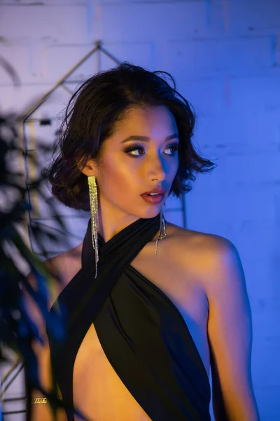 Confiado y elegante mujer asiática en vestido mirando lejos cerca de la planta en el club nocturno con luz de neón - foto de stock