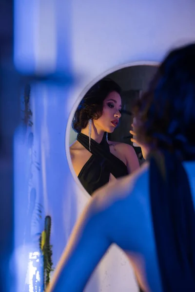 Стильная азиатка в платье делает селфи на смартфоне возле зеркала в ночном клубе с неоновым светом — стоковое фото