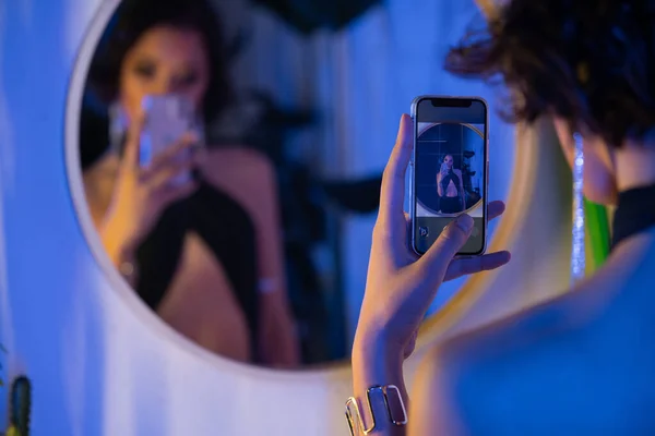 Размытая молодая азиатка в платье делает селфи на смартфоне возле зеркала в ночном клубе — стоковое фото