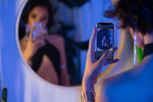 Blurred young asian woman in dress taking selfie on smartphone near mirror in night club — Stock Photo