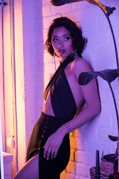 Привлекательная азиатка в платье с макияжем смотрит в сторону, стоя в неоновом свете в ночном клубе — стоковое фото