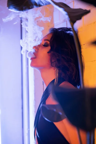 Trendy young brunette asian woman in dress exhaling smoke in neon light in night club — Stock Photo