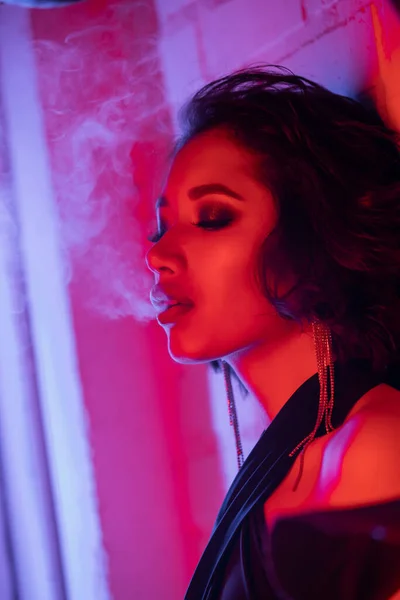 Moda joven asiático mujer con maquillaje exhalando humo mientras de pie en neón luz en noche club — Stock Photo