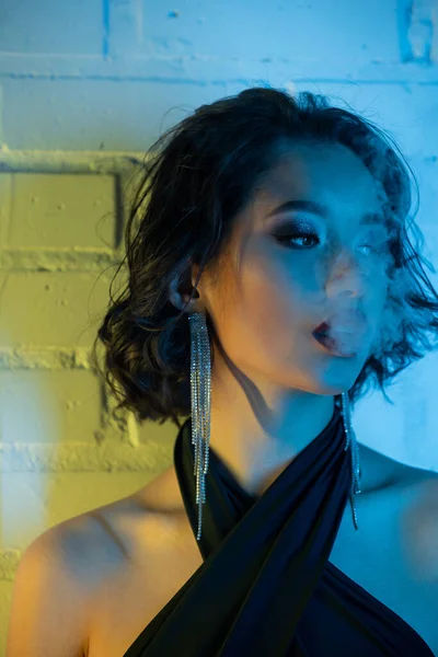 Retrato de mujer asiática joven de moda con maquillaje exhalando humo en luz de neón en club nocturno - foto de stock