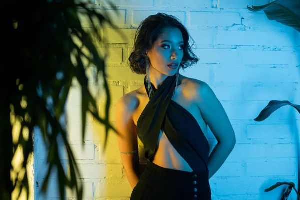 Alla moda giovane donna asiatica in abito in piedi vicino al muro e le piante in luce al neon in night club — Foto stock