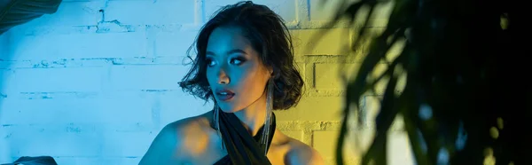 Elegante giovane donna asiatica guardando lontano e in piedi vicino a piante in luce al neon in night club, banner — Foto stock