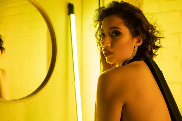 Na moda asiático mulher olhando para câmera enquanto em pé perto de espelho e fluorescente lâmpada no noite clube — Fotografia de Stock