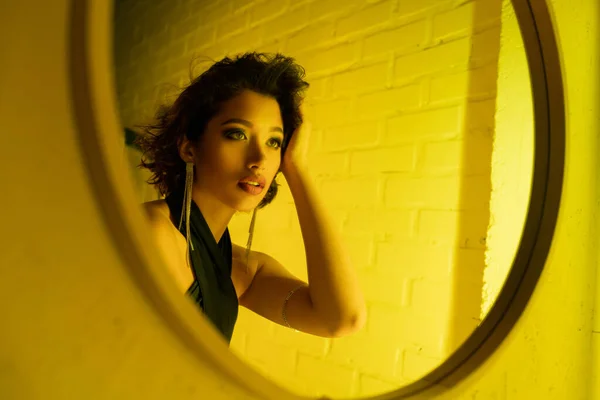 Elegante joven morena mujer asiática tocando el pelo y mirando el espejo en la luz de neón en el club nocturno - foto de stock