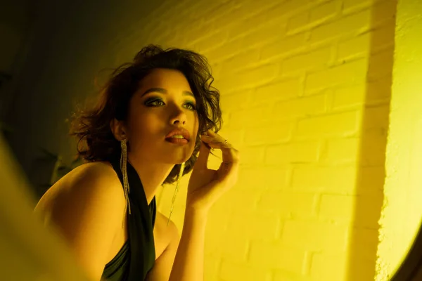 Joven mujer asiática en vestido elegante tocando el pelo cerca del espejo en el club nocturno con luz de neón - foto de stock
