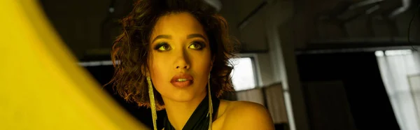 Fiduciosa ed elegante donna asiatica che guarda lo specchio in luce gialla nel night club, banner — Foto stock