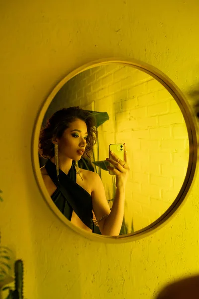 Elegante mujer asiática en vestido tomando selfie cerca de espejo e iluminación amarilla en club nocturno - foto de stock