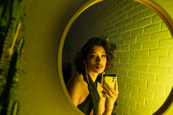 Asiatin im Kleid macht Selfie mit Smartphone in der Nähe von Spiegel und Pflanze in Nachtclub mit Neonlicht — Stockfoto