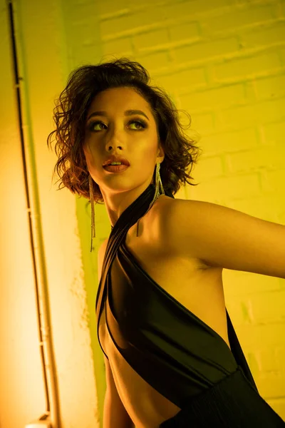 Retrato de jovem mulher asiática no vestido olhando embora enquanto estava em pé no clube noturno com luz de néon — Fotografia de Stock