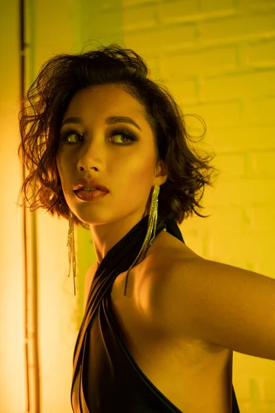 Junge asiatische Frau mit Ohrringen und Kleid schaut weg, während sie im Neonlicht in einem Nachtclub steht — Stockfoto