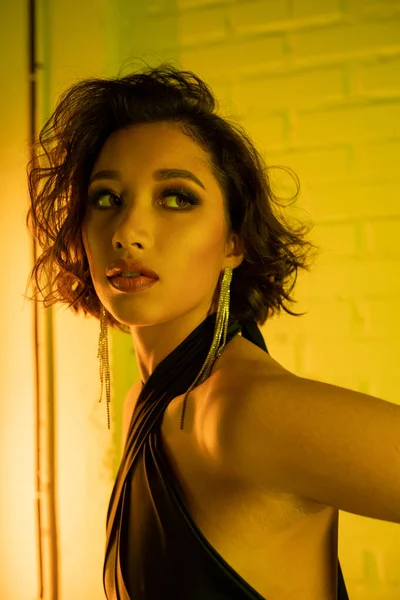 Young asian woman in earrings and dress looking away while standing in neon light in night club — Stock Photo