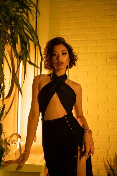 Sexy asiatische Frau im Kleid steht in der Nähe von Waschbecken und Pflanze in Nachtclub mit Neonbeleuchtung — Stockfoto