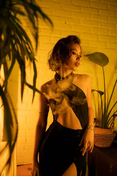 Sexy giovane donna asiatica in piedi vicino a piante in giallo illuminazione in night club — Foto stock