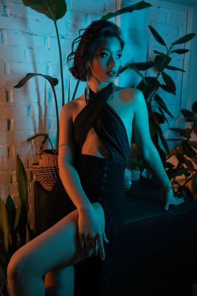 Elegante junge Asiatin im Kleid schaut in der Nähe von Pflanzen im Neonlicht in Nachtclub weg — Stockfoto