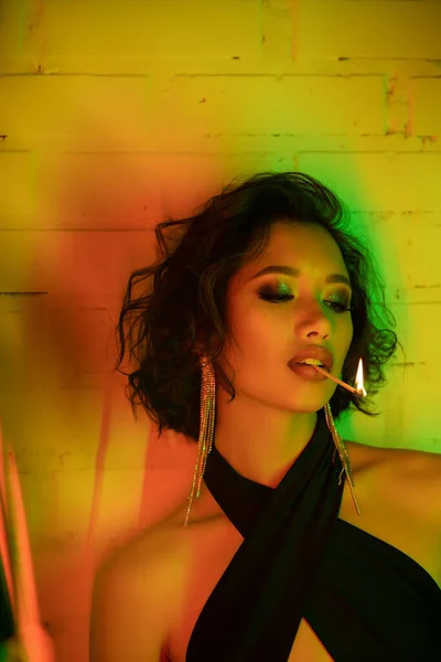 Asian woman in earrings and dress holding match with fire in lips in night club with neon light — Stock Photo