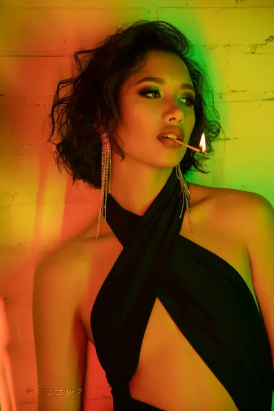 Sexy mujer asiática en vestido celebración partido con fuego en los labios, mientras que de pie en el club nocturno con iluminación - foto de stock