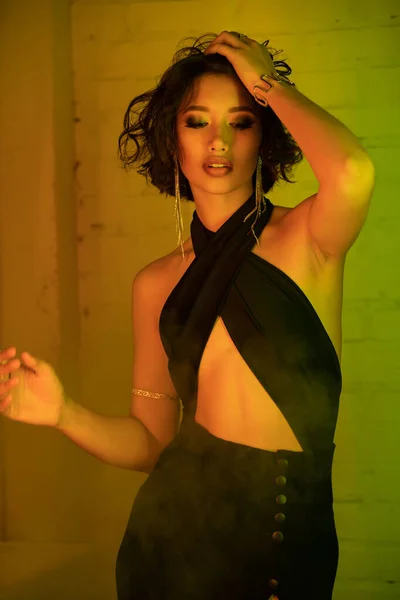 Sexy donna asiatica in abito toccare i capelli mentre in piedi in fumo e luce al neon in night club — Foto stock