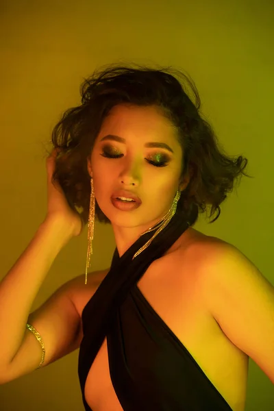 Elegante junge asiatische Frau mit Make-up im Kleid berühren Haare in Neonlicht in Nachtclub — Stockfoto