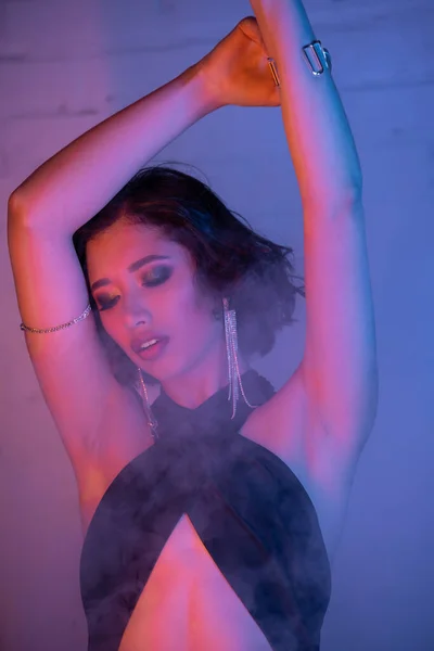 Модная молодая азиатка в платье танцует в дыму и неоновом свете в ночном клубе — стоковое фото