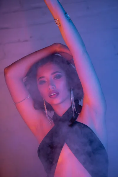 Trendige asiatische Frau in Kleid tanzt und schaut in Rauch und Neonlicht in Nachtclub in die Kamera — Stockfoto