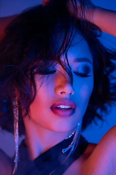 Elegante mujer asiática con peinado y maquillaje posando con los ojos cerrados en luz de neón en el club nocturno - foto de stock