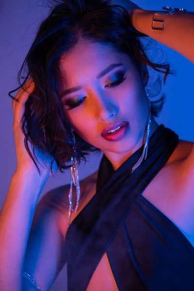 Portrait of young asian woman with makeup touching hair while posing in neon light in night club — Stock Photo